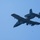 An A-10 Thunderbolt II (Warthog) ground-attack aircraft is photographed in flight against a clear blue sky. The aircraft appears to have just released a munition, which is visible falling below the fuselage. The image is taken from a ground-level perspective looking upward.