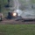 An armored military vehicle, partially concealed behind a earthen berm or hull-down position, is firing its main weapon, producing a visible muzzle flash and smoke. A secondary explosion or impact can be seen to the left near a utility pole, suggesting incoming fire or a simulated target detonation. The scene appears to be a military training exercise or live-fire demonstration set in a rural, green landscape.