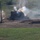 An armored military vehicle, partially concealed behind a earthen berm or sand mound, is firing its main weapon. A visible muzzle flash and multiple smoke plumes are present, indicating an active firing event. The scene appears to be a military training range or combat environment, with disturbed earth, dirt tracks, and green fields visible in the background.