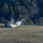 A blue vehicle, appearing to be a tractor or farm truck, is visible in an open field with a large white smoke or steam cloud billowing from it. Debris or liquid appears to be spraying outward from the vehicle, suggesting a mechanical failure, explosion, or pressurized release event. The scene is set against a backdrop of dense green and autumn-tinged trees.