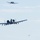 Three military aircraft, identifiable as A-10 Thunderbolt II (Warthog) ground-attack jets, are flying in a loose formation against a pale blue, lightly overcast sky. The foreground aircraft is clearly visible showing its distinctive twin turbofan engines mounted above the rear fuselage, straight wings, and underwing hardpoints with ordnance or equipment. Two additional aircraft are visible at greater distances in the upper center and lower right of the frame.
