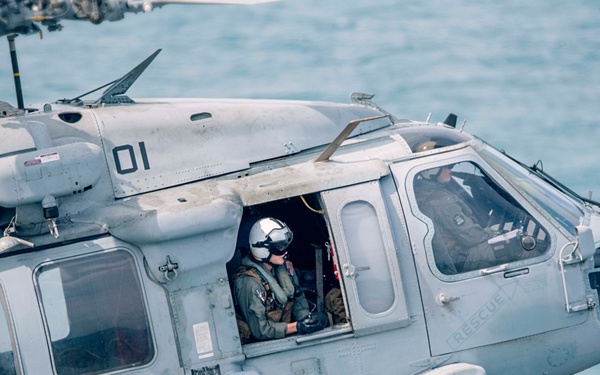 A U.S. Navy military helicopter, identified by the number '01' and an eagle insignia, is captured in flight over open water. The aircraft appears to be an MH-60S Knighthawk or similar Sikorsky Sea Hawk variant, with multiple crew members visible through the windows and open door. The helicopter is equipped with a rescue hoist and sensor pod, indicating a search and rescue or utility mission role.