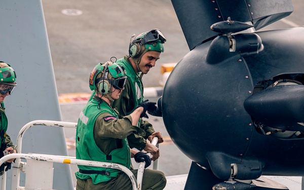Three military aviation technicians wearing green flight suits and helmets with hearing protection are working on an aircraft engine/propeller assembly on what appears to be a ship's flight deck. They are standing on a maintenance platform or work stand labeled 'GLBW 5788-001'. The personnel appear to be conducting inspection or maintenance on a large turboprop or propeller-driven aircraft.