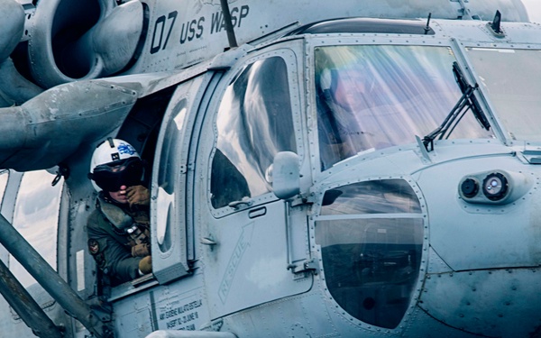 A U.S. Navy helicopter, marked '07' and assigned to USS WASP, is positioned on what appears to be a ship's flight deck over open water. A crew member in a flight helmet and gear is visible leaning out of the open side door. The aircraft appears to be an MH-60 Seahawk variant in standard naval gray livery.