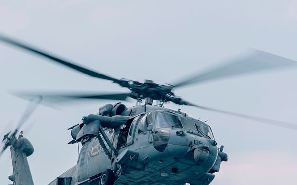 A military helicopter, consistent with a Sikorsky MH-60 Seahawk variant, is captured in flight against an overcast sky. The aircraft is painted in a standard naval gray scheme and appears to be in a hover or low-speed flight configuration. Rotor blades show motion blur indicating active flight.