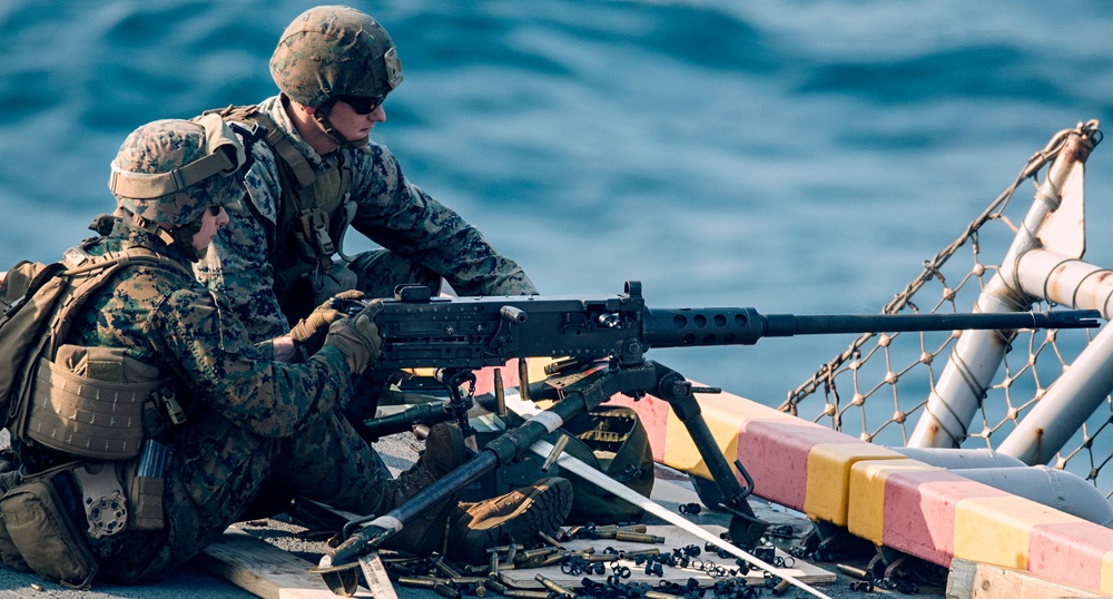 Two military personnel in MARPAT camouflage uniforms are positioned on what appears to be a naval vessel deck, operating a heavy machine gun. One service member is in a prone/kneeling firing position while the other appears to be assisting or instructing. Spent brass casings and weapon components are scattered on the deck surface around them.