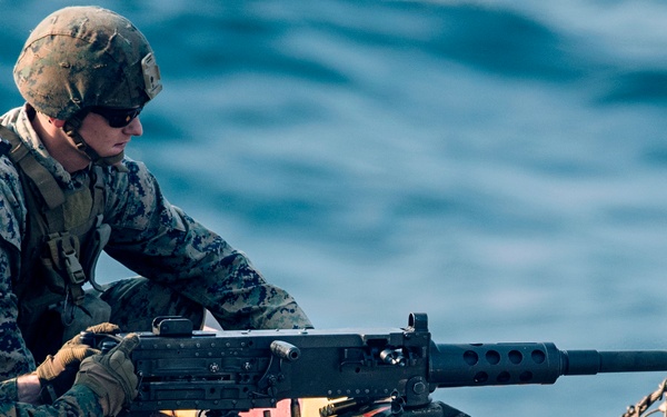 Two military personnel in MARPAT camouflage uniforms are positioned on what appears to be a naval vessel deck, operating a heavy machine gun. One service member is in a prone/kneeling firing position while the other appears to be assisting or instructing. Spent brass casings and weapon components are scattered on the deck surface around them.