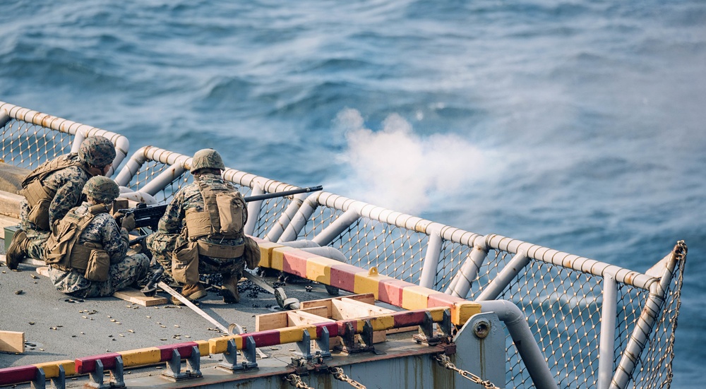 Several military personnel in camouflage uniforms are positioned on the deck of a naval vessel, firing a weapon over the side railing toward open water. A visible plume of smoke or muzzle blast is seen near the barrel of the weapon being discharged. The scene appears to be a live-fire training exercise conducted at sea.