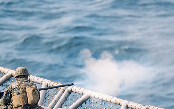 Several military personnel in camouflage uniforms are positioned on the deck of a naval vessel, firing a weapon over the side railing toward open water. A visible plume of smoke or muzzle blast is seen near the barrel of the weapon being discharged. The scene appears to be a live-fire training exercise conducted at sea.