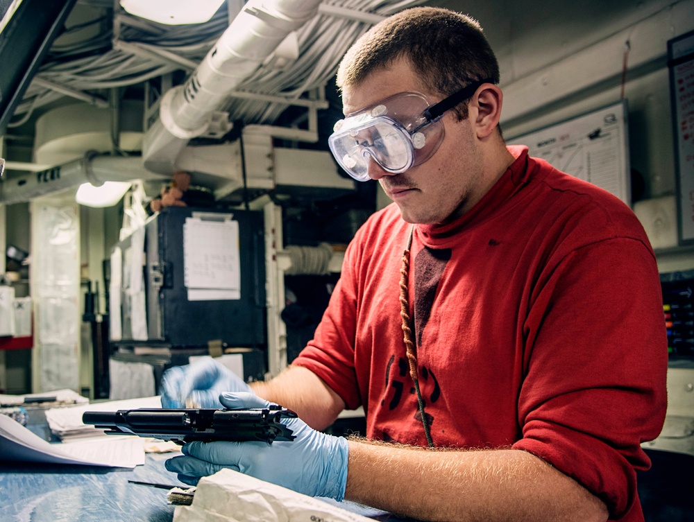 A young man wearing safety goggles and blue nitrile gloves is working on what appears to be a firearm, likely performing cleaning or maintenance. He is dressed in a red sweatshirt and is seated at a workbench in what appears to be an industrial or military workspace. The environment includes overhead piping, paperwork on walls, and various tools on the work surface.
