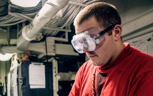 A young man wearing safety goggles and blue nitrile gloves is working on what appears to be a firearm, likely performing cleaning or maintenance. He is dressed in a red sweatshirt and is seated at a workbench in what appears to be an industrial or military workspace. The environment includes overhead piping, paperwork on walls, and various tools on the work surface.