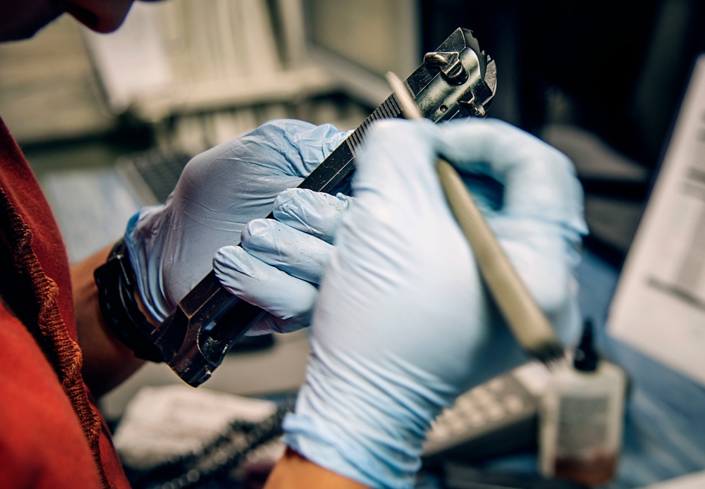 A person wearing blue nitrile gloves is handling a disassembled semi-automatic pistol, applying what appears to be a tool or brush to the firearm's components. The setting appears to be a workbench environment, consistent with firearm maintenance, forensic examination, or evidence processing. Various tools and containers are visible in the background.