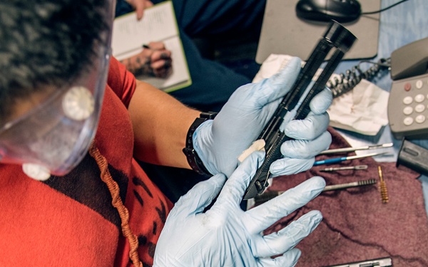 A technician wearing blue latex gloves and a face shield is examining or cleaning a disassembled firearm on a work surface. The workspace contains gun components, cleaning tools, and a landline telephone, suggesting a professional firearms maintenance or forensic examination setting. A second person is visible in the background taking notes.
