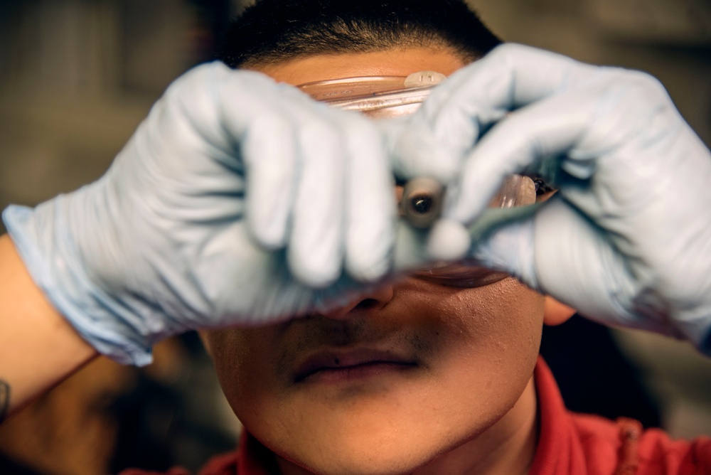 A person wearing blue latex gloves is holding a transparent object, likely a petri dish or glass slide, up close to their face and examining it carefully. The subject is wearing a red shirt and appears to be in a laboratory or workshop setting. The focus is on the gloved hands and the object being inspected, with the face slightly out of focus.