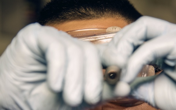 A person wearing blue latex gloves is holding a transparent object, likely a petri dish or glass slide, up close to their face and examining it carefully. The subject is wearing a red shirt and appears to be in a laboratory or workshop setting. The focus is on the gloved hands and the object being inspected, with the face slightly out of focus.