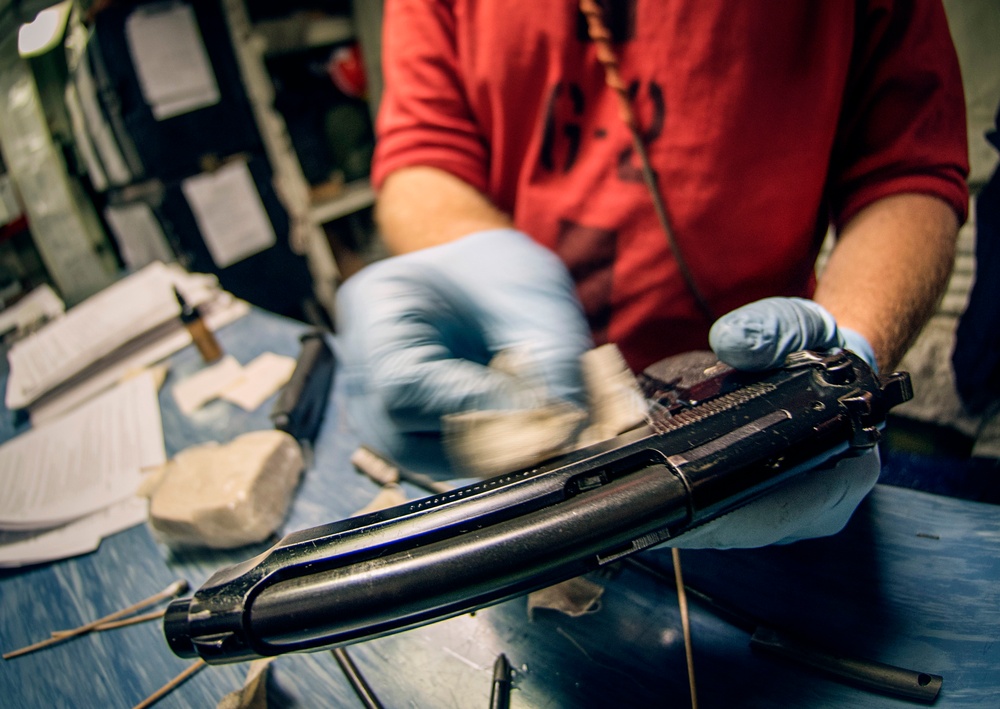 A person wearing blue latex gloves is cleaning or restoring a disassembled firearm on a blue work surface. The firearm appears to be a pistol or submachine gun with its slide or receiver separated, being worked on with cloths or cleaning patches. The workspace contains various tools, papers, and cleaning materials consistent with a gunsmithing or conservation environment.