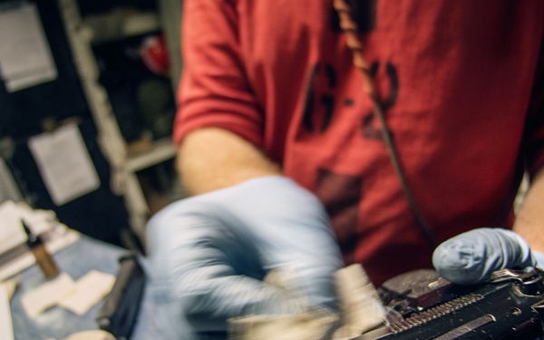A person wearing blue latex gloves is cleaning or restoring a disassembled firearm on a blue work surface. The firearm appears to be a pistol or submachine gun with its slide or receiver separated, being worked on with cloths or cleaning patches. The workspace contains various tools, papers, and cleaning materials consistent with a gunsmithing or conservation environment.