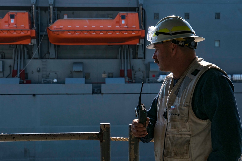 A male maritime or dock worker stands on what appears to be a vessel or dock structure, holding a two-way radio and looking to the left. He is wearing standard maritime safety gear including a hard hat with safety goggles, a safety vest labeled 'STEARNS', and a dark long-sleeve shirt. In the background, two orange enclosed lifeboats are mounted on davits on the side of a large vessel.