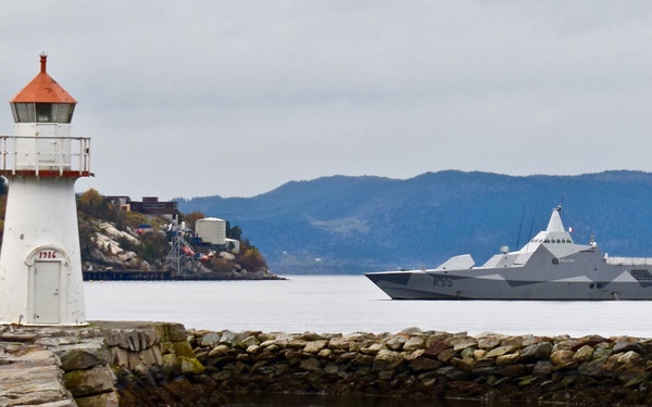A small white lighthouse with a red-orange cap, dated 1916, stands on a stone breakwater in the foreground. In the background, a modern stealth-design naval warship bearing the hull number 'K33' passes through calm fjord waters. The scene is set against a backdrop of forested hills and an overcast sky, consistent with a Scandinavian coastal environment.