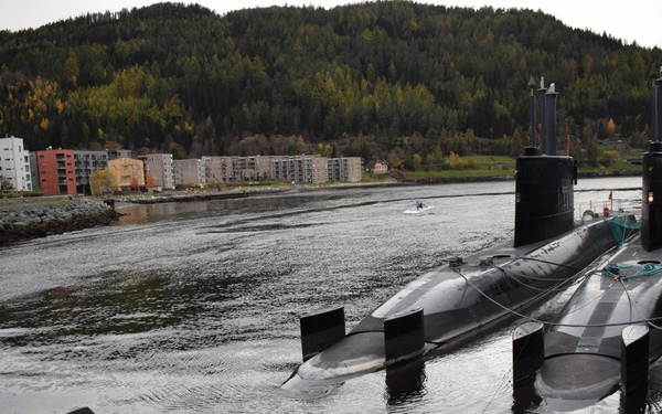 A dark-colored submarine is moored alongside a dock or quay in a calm harbor or fjord setting. Several of the submarine's control surfaces (fins/hydroplanes) are visible above the waterline, with some appearing partially submerged. The background features residential buildings and a forested hillside, suggesting a Scandinavian or Northern European location.