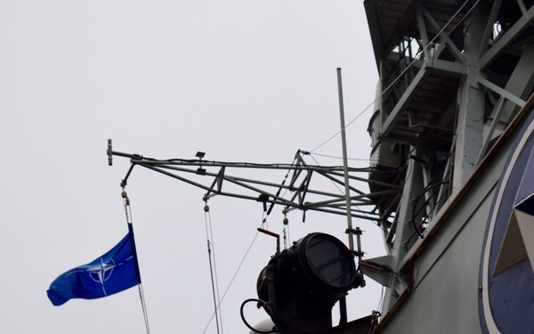 The image shows the upper superstructure of a naval vessel flying a NATO flag. A large searchlight/signal lamp is mounted on the mast structure, and a circular emblem featuring a dark blue ring with a triangular arrow symbol is visible on the ship's hull. The overcast sky provides a grey backdrop.