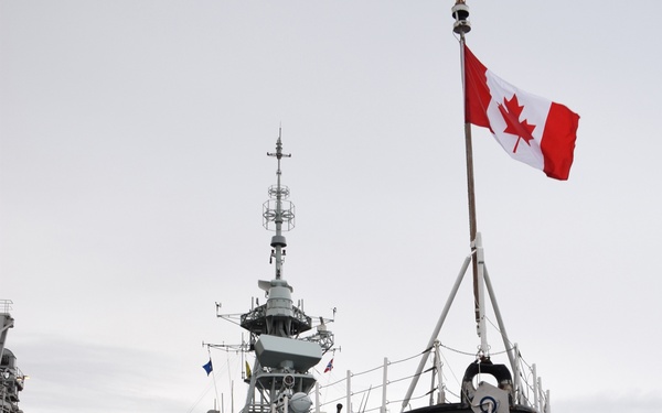 A grey Canadian naval warship is moored at a dock, viewed from a bow-on perspective. The Canadian flag (Maple Leaf) flies prominently from a flagstaff at the bow. An orange oil containment boom surrounds the hull at the waterline, suggesting maintenance or environmental precaution activity.