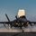 First test bombs dropped from HMS Queen Elizabeth’s F-35 Lightning fighter jets A fifth-generation stealth fighter jet, identifiable as an F-35B, is captured during a short takeoff or landing on what appears to be an aircraft carrier deck. The lift fan door is open on top of the fuselage, characteristic of the F-35B STOVL variant. Steam or exhaust vapor billows dramatically around the aircraft, suggesting catapult steam or engine exhaust during the operation.