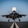 First test bombs dropped from HMS Queen Elizabeth’s F-35 Lightning fighter jets A front-facing view of an F-35B Lightning II stealth fighter jet captured during a short takeoff or vertical landing maneuver on a runway. The aircraft's landing gear is fully deployed and a lift fan door is open on top of the fuselage, characteristic of the F-35B STOVL variant. The image is taken from a low, head-on perspective, emphasizing the aircraft's imposing silhouette against a clear blue sky.