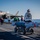 First test bombs dropped from HMS Queen Elizabeth’s F-35 Lightning fighter jets The image shows flight deck operations aboard a U.S. Navy aircraft carrier, with an F-35C Lightning II stealth fighter jet visible in the background. In the foreground, a weapons cart carrying what appears to be air-to-air or air-to-ground munitions with red safety streamers is being managed by deck crew. Personnel wearing color-coded jerseys and helmets are conducting various flight deck duties under clear skies over open ocean.