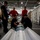 First test bombs dropped from HMS Queen Elizabeth’s F-35 Lightning fighter jets Military personnel are gathered in what appears to be a naval vessel's hangar bay, working on a munition or ordnance item resting on a cart or dolly. Five individuals are present, with two in red jerseys (typically indicating ordnance handlers on US Navy vessels) and others in dark navy uniforms. The setting includes shipping containers along the right side and aircraft visible in the background.
