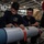FIRST BOMBS DROPPED FROM HMS QUEEN ELIZABETH’S F-35 FIGHTER JETS Two naval personnel in dark blue uniforms are working on what appears to be an air-launched weapon or munition in what looks like a ship's hangar bay. The individual on the left wears a patch identifying him as 'BARRAS' from '815 Naval Air Squadron.' A red 'REMOVE BEFORE FLIGHT' safety tag is visible on the munition being handled.