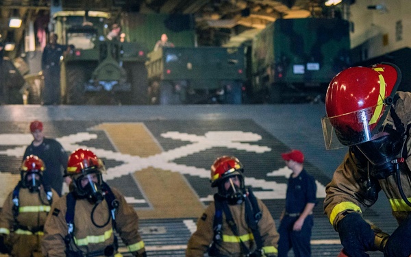 A group of personnel in full firefighting gear, including tan proximity suits, red helmets, and SCBA breathing apparatus, are conducting a firefighting drill aboard what appears to be a military vessel's well deck or hangar bay. The foreground figure is crouching while managing a red fire hose, while three additional suited personnel advance in formation behind. Military vehicles, including what appear to be armored trucks, are visible in the background along with observers in standard uniforms.
