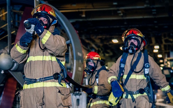 The image shows a group of firefighters in full protective gear, including turnout suits, helmets, and SCBA (Self-Contained Breathing Apparatus) masks, operating inside what appears to be a naval vessel's hangar bay. They are advancing with a fire hose in a coordinated team formation, suggesting a firefighting drill or actual emergency response. A large aircraft propeller is visible in the background, confirming the naval/aviation environment.
