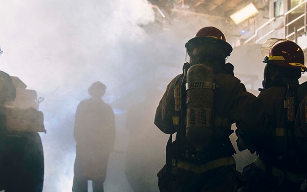 The image depicts firefighters in full protective gear, including SCBA (Self-Contained Breathing Apparatus) units branded 'Scott', advancing through a smoke-filled environment. Several silhouetted figures are visible in the background amid heavy white smoke. The setting appears to be a large industrial or hangar-type structure, possibly a training exercise or active emergency response.