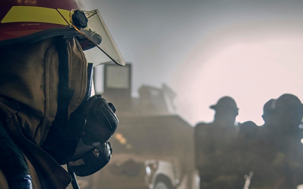 The image depicts firefighters operating in a smoke-filled environment, with one firefighter prominently in the foreground wearing full protective gear including a helmet, face mask/respirator, and turnout coat. Several other firefighters are visible in the background as silhouettes against a bright, hazy light source. A large vehicle, likely a fire truck or military-style vehicle, is partially visible in the mid-ground.