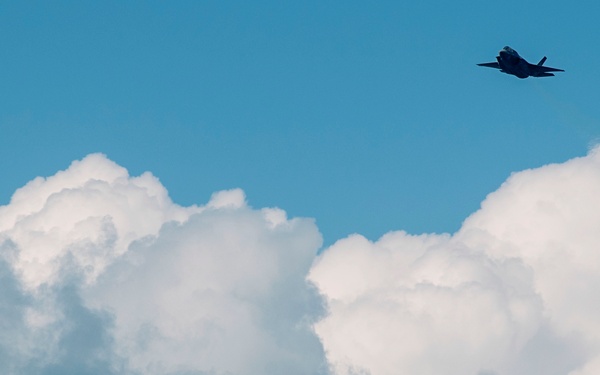 Two military jet aircraft are visible flying through a partly cloudy blue sky. The upper-right aircraft is clearly silhouetted and appears to be a fifth-generation stealth fighter, likely an F-35, while a second aircraft is visible in the lower-left at greater distance. The scene appears to be captured during daylight with large cumulus clouds forming a dramatic backdrop.