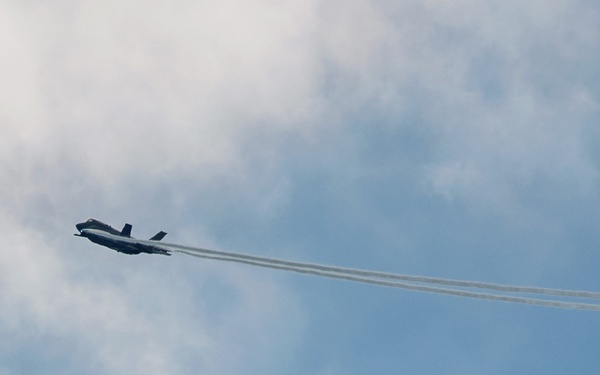 A military jet aircraft is captured in flight against a partly cloudy blue sky, leaving two distinct condensation trails (contrails) behind it. The aircraft appears to be banking or maneuvering, photographed from a ground-level upward angle. The contrails extend diagonally across the frame from upper-left to lower-right.