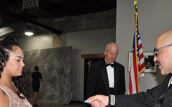 A U.S. Navy sailor in dress uniform is handing something to a young woman in a sequined dress at a registration or check-in table during a formal military event. An older gentleman in a tuxedo stands in the background near an American flag. The setting appears to be a banquet hall or event venue.