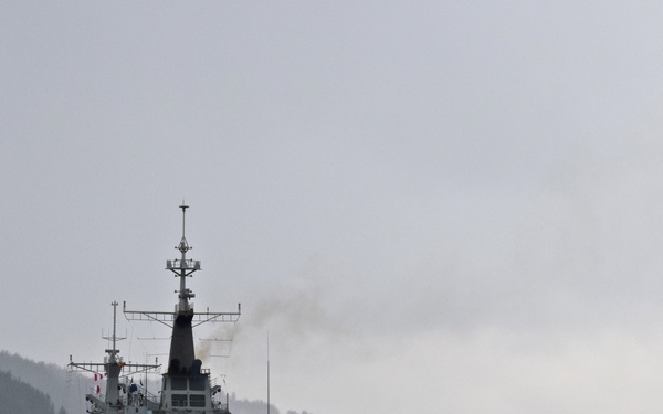 A naval vessel with hull number 'A15' is underway on a calm body of water, surrounded by misty, forested hills in the background. The ship appears to be a military auxiliary or support vessel, painted in standard naval gray. Light exhaust smoke is visible rising from the funnel, indicating active propulsion.