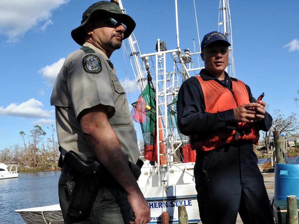 Two uniformed officials stand on a dock near a white fishing vessel named 'Southern Style' from what appears to be a Florida port. One officer wears a Florida Fish and Wildlife Conservation Commission uniform with a wide-brim hat and holstered sidearm, while the other wears a U.S. Coast Guard cap and orange life vest. The surrounding area shows storm-damaged trees, suggesting this is a post-hurricane response or assessment operation.