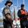 Two uniformed officials stand on a dock near a white fishing vessel named 'Southern Style' from what appears to be a Florida port. One officer wears a Florida Fish and Wildlife Conservation Commission uniform with a wide-brim hat and holstered sidearm, while the other wears a U.S. Coast Guard cap and orange life vest. The surrounding area shows storm-damaged trees, suggesting this is a post-hurricane response or assessment operation.