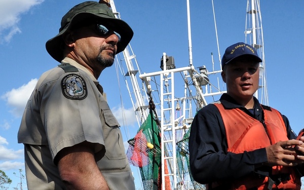 Two uniformed officials stand on a dock near a white fishing vessel named 'Southern Style' from what appears to be a Florida port. One officer wears a Florida Fish and Wildlife Conservation Commission uniform with a wide-brim hat and holstered sidearm, while the other wears a U.S. Coast Guard cap and orange life vest. The surrounding area shows storm-damaged trees, suggesting this is a post-hurricane response or assessment operation.