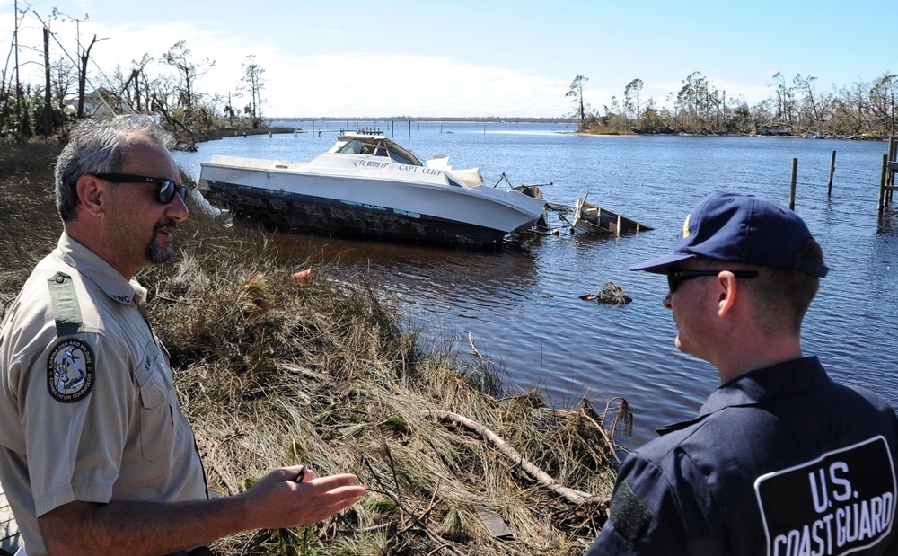 A U.S. Coast Guard officer and a Florida Fish and Wildlife Conservation Commission (FWC) officer survey storm damage along a waterway. A partially sunken or grounded motorboat named 'CAPT. CLIFF' with Florida registration is visible in the background, along with debris and damaged trees consistent with hurricane aftermath. The scene appears to be a post-storm damage assessment operation.