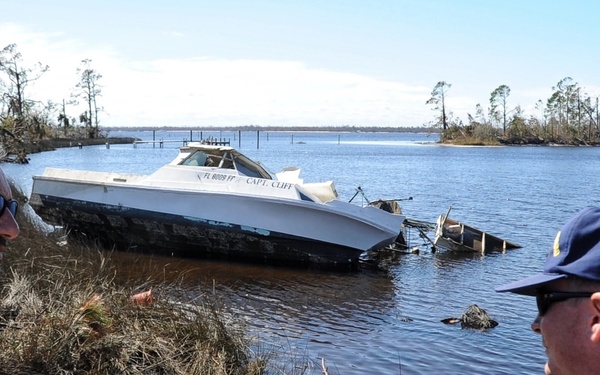 A U.S. Coast Guard officer and a Florida Fish and Wildlife Conservation Commission (FWC) officer survey storm damage along a waterway. A partially sunken or grounded motorboat named 'CAPT. CLIFF' with Florida registration is visible in the background, along with debris and damaged trees consistent with hurricane aftermath. The scene appears to be a post-storm damage assessment operation.