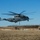 A U.S. Marines CH-53E Super Stallion heavy-lift helicopter, marked with the number '04' and 'MARINES' on the fuselage, is hovering at low altitude over a cleared field area. The helicopter appears to be conducting a sling-load or external lift operation, with a hook/line visible beneath the aircraft. Traffic cones mark a designated landing or operations zone on a paved surface nearby, with a military ground vehicle and personnel visible in the background.