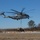 A U.S. Marines CH-53K King Stallion heavy-lift helicopter (tail number 04) is performing a sling-load operation, lifting a military tactical vehicle from a marked landing zone. Several ground personnel are visible managing the rigging and load. The scene appears to be a training exercise conducted at a military airfield or training area during winter conditions.
