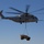 A U.S. Marines heavy-lift helicopter, marked '04' and labeled 'MARINES', is airborne and conducting an external sling-load operation. It is carrying a tan/desert-colored military tactical vehicle suspended beneath it via sling rigging. The sky is clear and blue, suggesting daytime operations in a dry or arid environment.