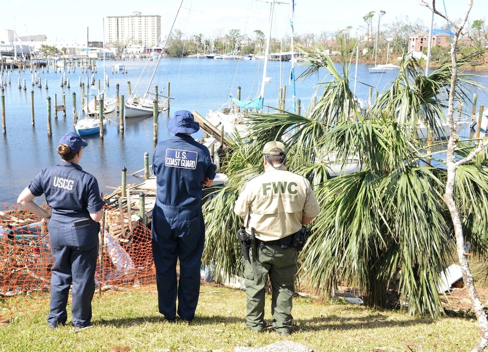 Three law enforcement officers — two U.S. Coast Guard (USCG) personnel and one Florida Fish and Wildlife Conservation Commission (FWC) Law Enforcement officer — are surveying storm damage at a marina. The scene shows significant debris, displaced boats, damaged docks, and downed palm trees consistent with hurricane or major storm aftermath. The officers appear to be conducting a post-storm damage assessment along a coastal waterway.