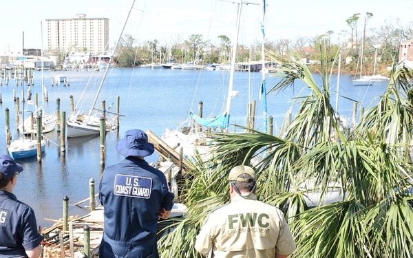 Three law enforcement officers — two U.S. Coast Guard (USCG) personnel and one Florida Fish and Wildlife Conservation Commission (FWC) Law Enforcement officer — are surveying storm damage at a marina. The scene shows significant debris, displaced boats, damaged docks, and downed palm trees consistent with hurricane or major storm aftermath. The officers appear to be conducting a post-storm damage assessment along a coastal waterway.