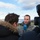 An outdoor press interview is taking place near a body of water, likely a fjord or lake. A person in military camouflage uniform is being interviewed by a journalist holding an NRK (Norwegian Broadcasting Corporation) microphone. A camera operator is filming the exchange from the right side of the frame.